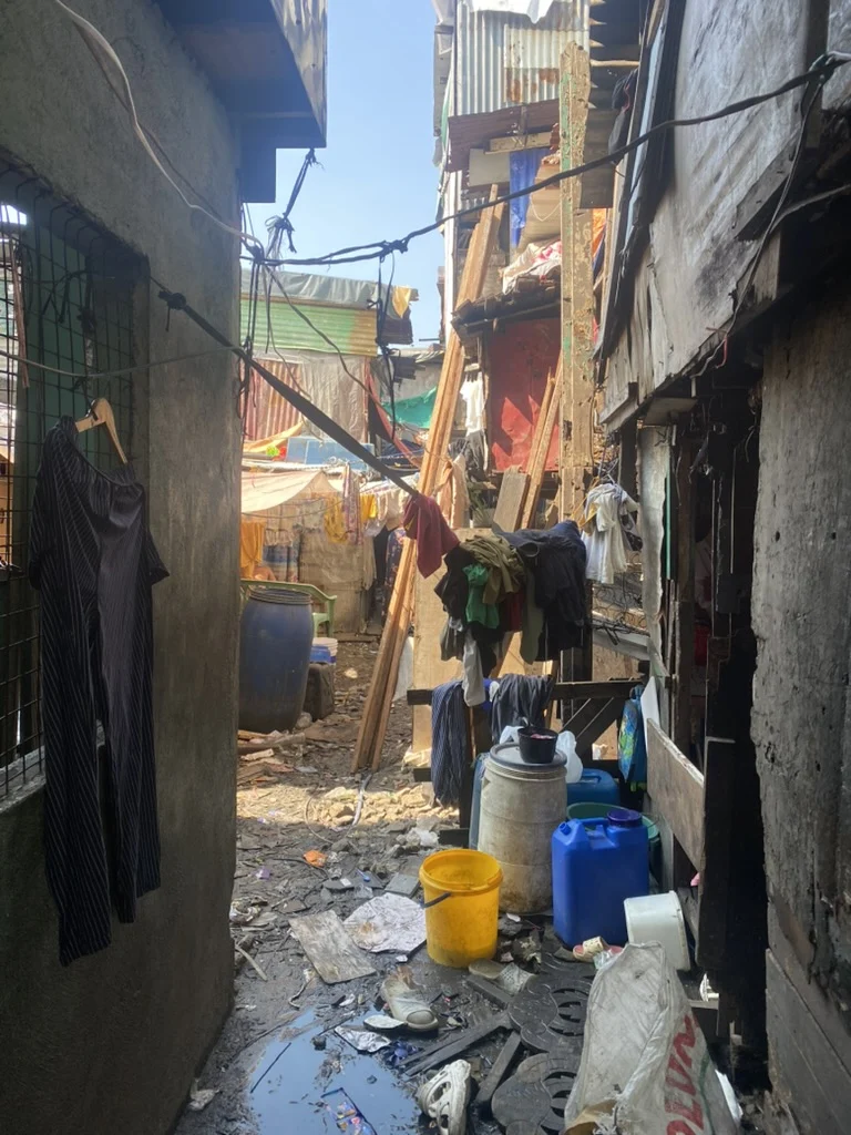 A narrow lane with uneven ground, containers and scattered rubbish.